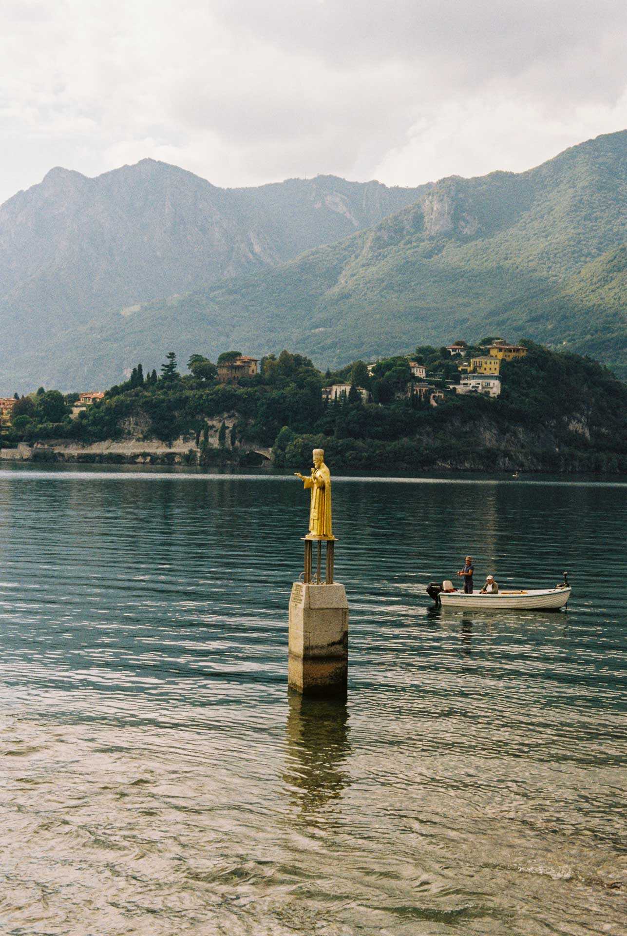 A gold statue of the Pope stands tall in the water beside a fishing boat with two men fishing the waters at Lecco, Italy, with the mountains behind them. Taken on Kodak Gold 200 film.