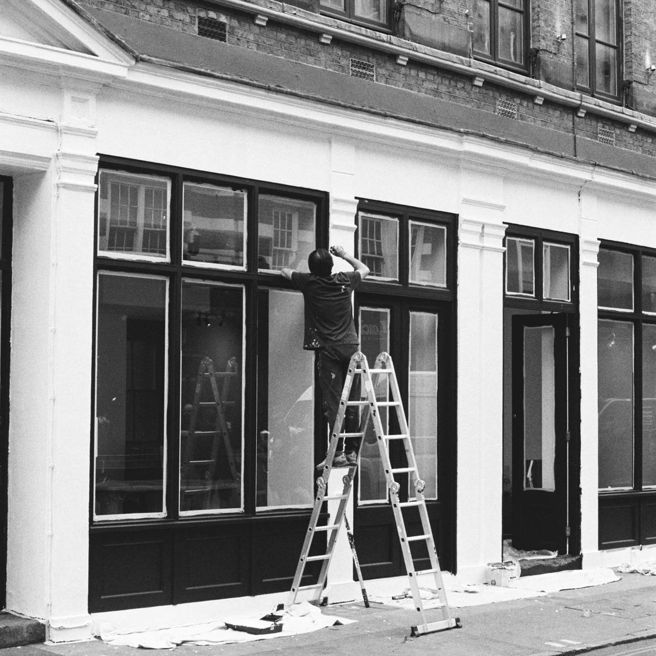 A black and white photograph taken on Ilford HP5 Plus film stock showing a man climbing a ladder in London.