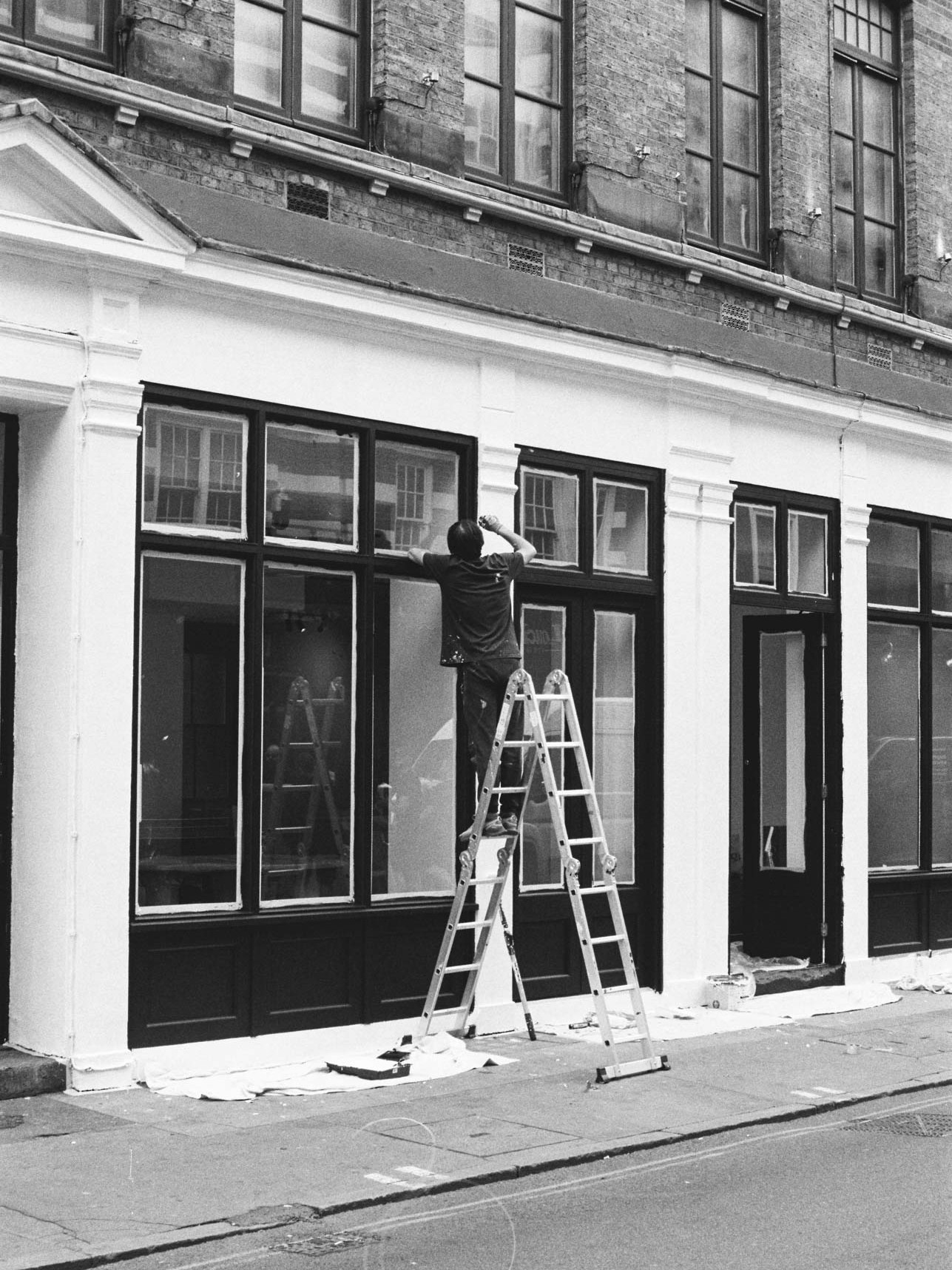 A black and white photograph taken on Ilford HP5 Plus film stock showing a man climbing a ladder in London.